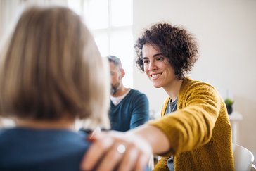 Eine lächelnde Frau ist im Gespräch mit einer Person und legt ihr beruhigend die Hand auf die Schulter.
