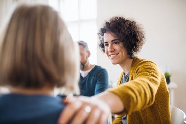 Eine lächelnde Frau ist im Gespräch mit einer Person und legt ihr beruhigend die Hand auf die Schulter.
